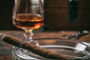 A fine cigar rests in a glass ashtray with a glass of brandy in the soft focus background.